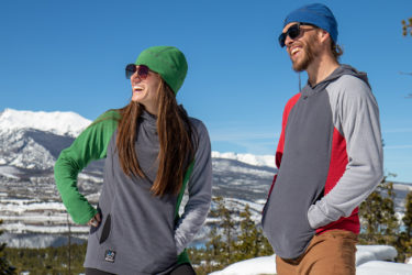 A woman and man in hoodies and beanies stand on top of a mountain in winter