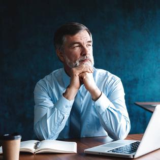 Focused older businessman deep in thought while sitting at a table in modern office with a laptop and a notebook in front of him.