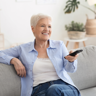 An older woman sits on the sofa and flips through the TV channels with a remote.