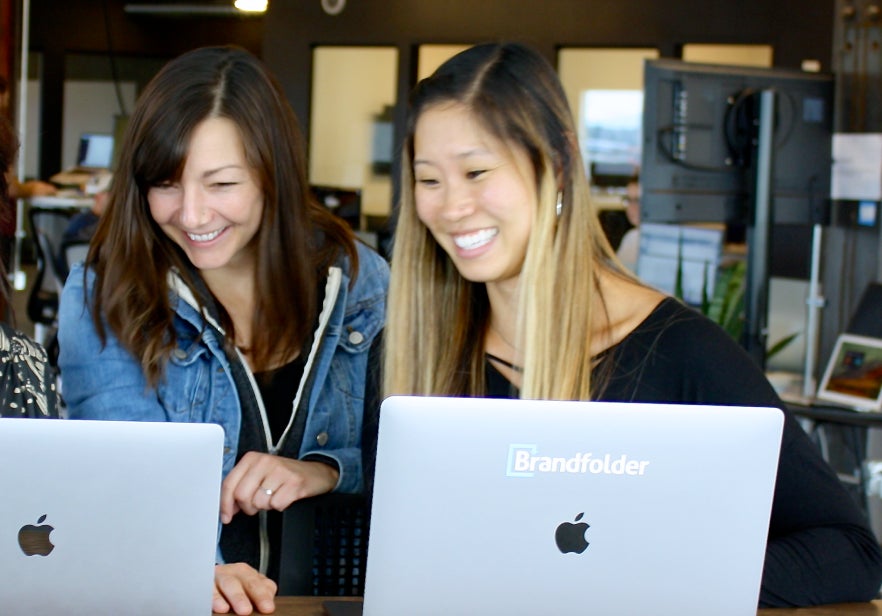 Two smiling women with Brandfolder laptops look at one of the laptop screens.