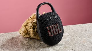 The JBL Clip 5 in black is pictured leaning against a sand colored rock against a pink background,