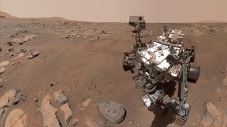 NASA's Perseverance rover to the right of the image took this selfie amidst the barren, dusty and rocky Martian landscape. Some larger rocks can be seen in the foreground and background and the sky is a hazy red color.