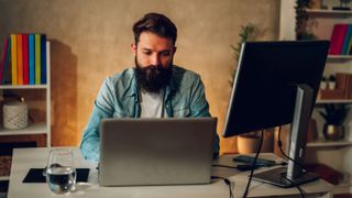 A bearded man sitting at a table that has a laptop and a second monitor