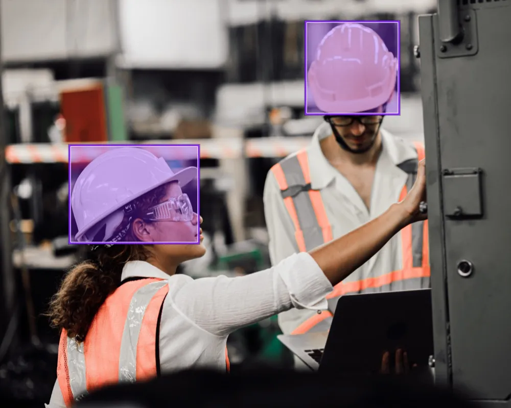 Two manufacturing workers wearing PPE, with bounding boxes around their hard hats 