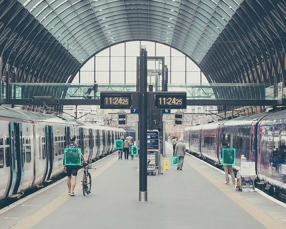 Two trains at a train station, with people walking down the platform, and bounding boxes drawn around bags. 