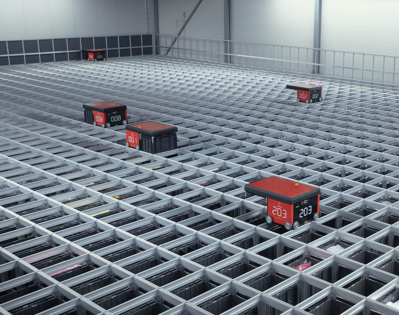 An AutoStore grid viewed from above. Four AutoStore R5 robots at work. One R5 robot in the left hand corner positioned at a charging port. Photo. 