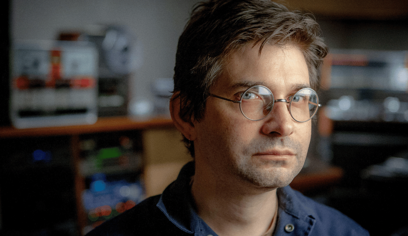 S: a man in his 40s, with brown hair and round glasses, in his studio. 