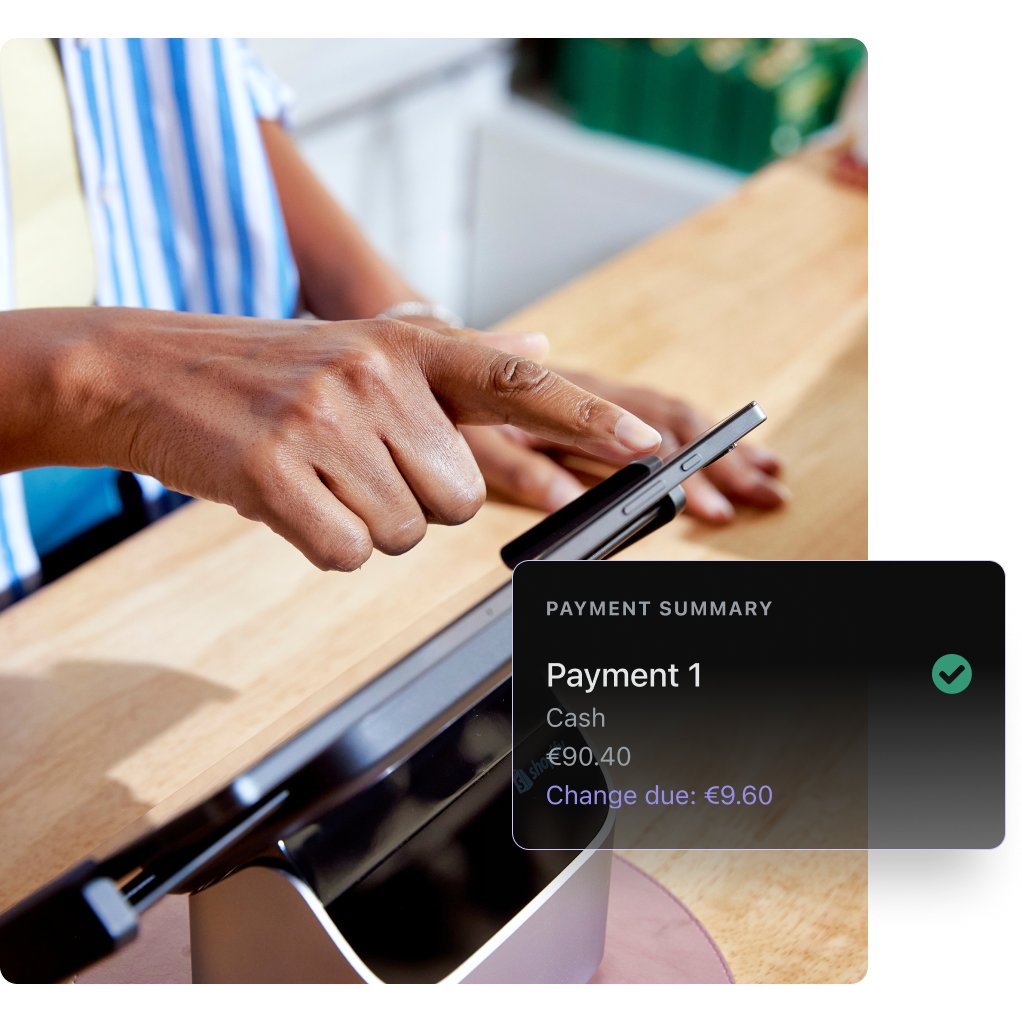 A closeup of a checkout counter where an associate where a blue and white striped overshirt is typing on a POS tablet. The tablet and stand are on a pale wood counter. Overlaid on the image is a Shopify POS UI screen indicating a payment summary.