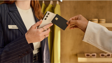 Close up of a customer using their credit card to make a purchase with Tap to Pay on Android in a large retail environment. A sales associate in a navy pinstriped blazer holds the Android device, while the customer offers a credit card to tap. The background shows shelves stocked with neatly arranged accessories.