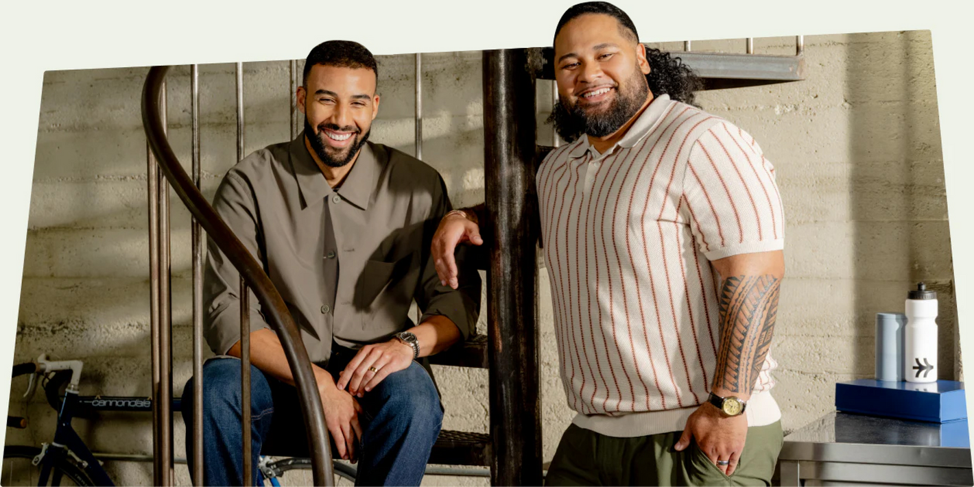 Two Shopify merchants smiling on a spiral staircase. One wears a gray shirt and jeans, while the other is in a striped polo and green pants. A bicycle is in the background, and a table holds a water bottle and a blue box.