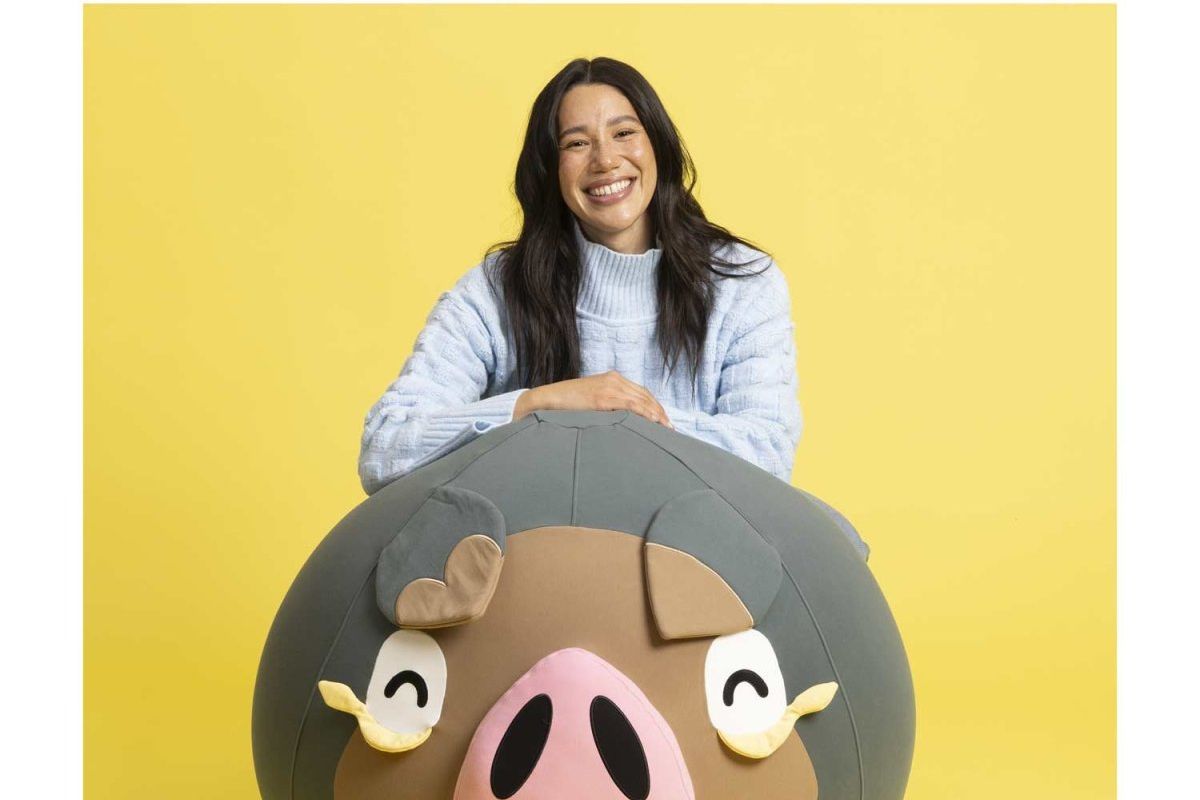 A photo of a person sitting behind a big round Lechonk beanbag. They’re smiling and there is a yellow background. The Lechonk looks really big and round.