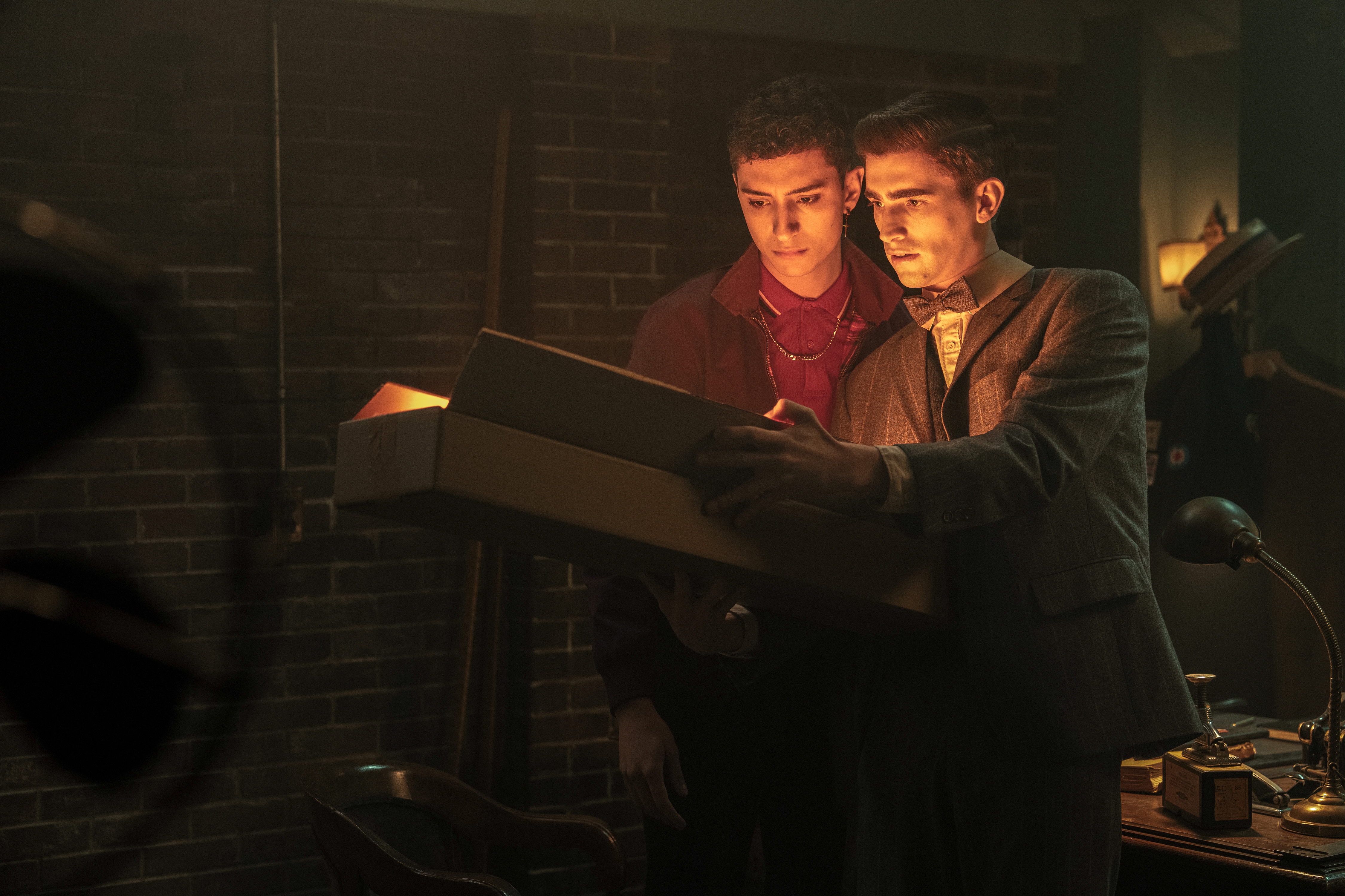 (L to R) Jayden Revri as Charles Rowland and George Rexstrew as Edwin Payne in Dead Boy Detectives. They’re opening a big box, with a light from within illuminating their faces. 