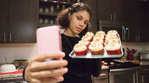 Girl holding up a tray of cupcakes in front of a cellphone