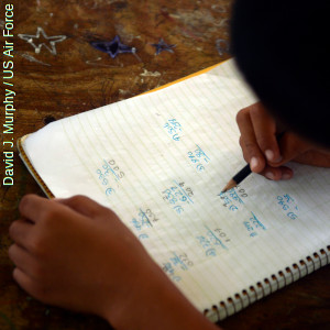 Child's hand writing maths classwork in an open lined notebook.