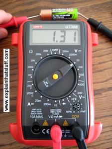 A digital voltmeter standing with red and black probes attached, wired into a circuit to test a simple 1.5-volt battery.
