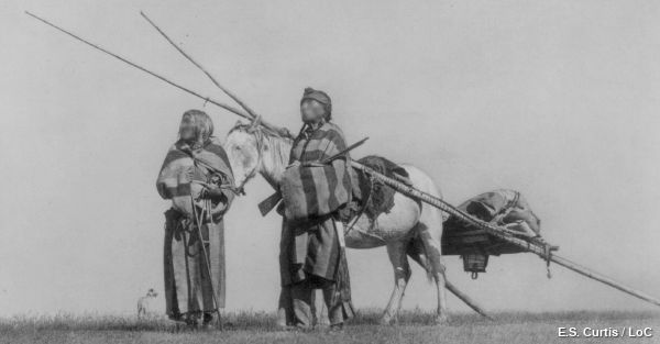 Two Native Americans walk alongside a horse and travois.
