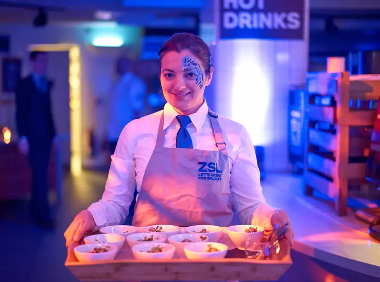 A young woman with facepaint on the left side of her face holding a tray of bowl food at an evening event