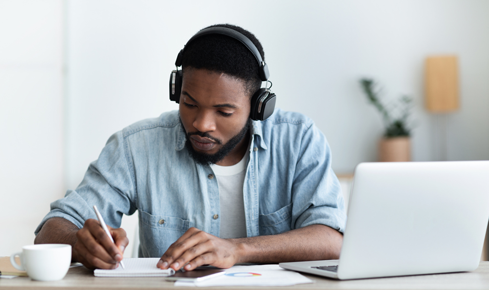 молодой темнокожий мужчина в наушниках пишет заметки, сидя за столом с ноутбуком