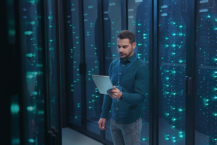 IT professional standing in server room with tablet, monitoring system performance of database