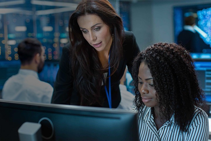 Female Team Leader Consults Young Computer Engineer. They Work in a Crowded Office on a Neural Network/ Artificial Intelligence Project.