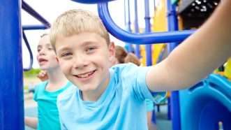 A child smiles into the camera as other kids play on the playground behind him | Yuri Arcurs | Dreamstime.com