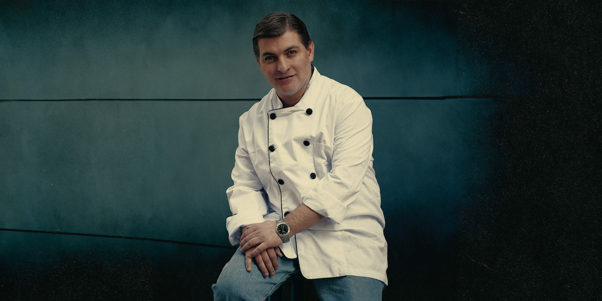 César Román wears a chef’s jacket and sits against a grey-green background in an archival photo from the docuseries ‘Cooking Up Murder: Uncovering the Story of César Román’