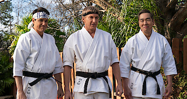 Ralph Macchio as Daniel LaRusso, William Zabka as Johnny Lawrence and Yuji Okumoto as Chozen stand wearing white karate uniforms with black belts in Season 6 of 'Cobra Kai'