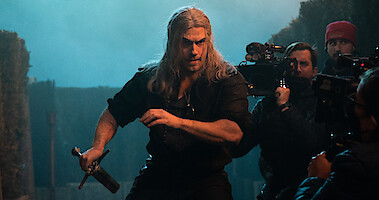 Henry Cavill as Geralt of Rivia in Season 3 of 'The Witcher' stands with his sword drawn while members of the film crew stand around him holding cameras. 