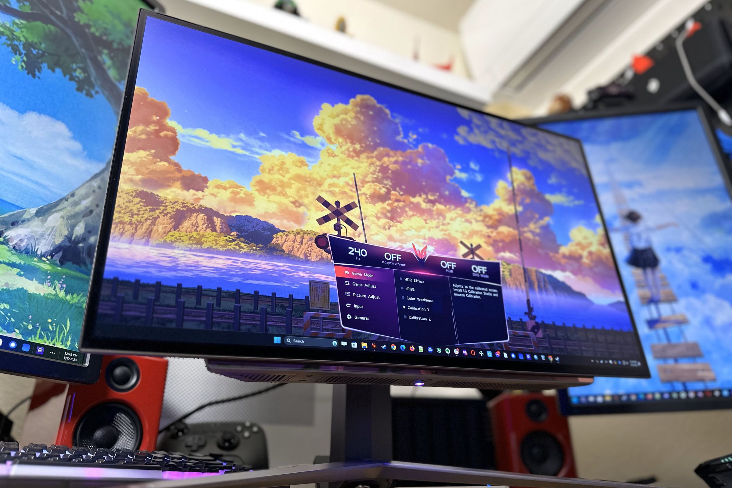 A bright, colorful OLED monitor with a big V-shaped stand underneath, flanked by two other monitors in portrait mode. The main monitor has a colorful sky on screen with big orange sunset-lit clouds and an on-screen display showing it’s running at 240Hz.