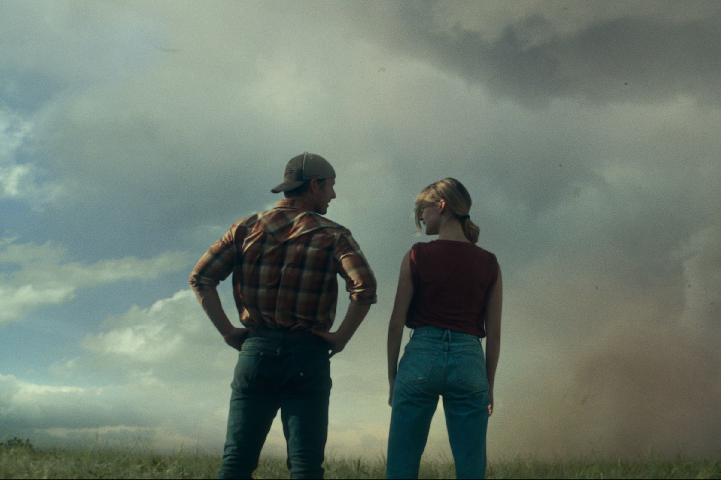 A woman and man standing side by side in a wide open field as they face a massive tornado twisting in the distance.