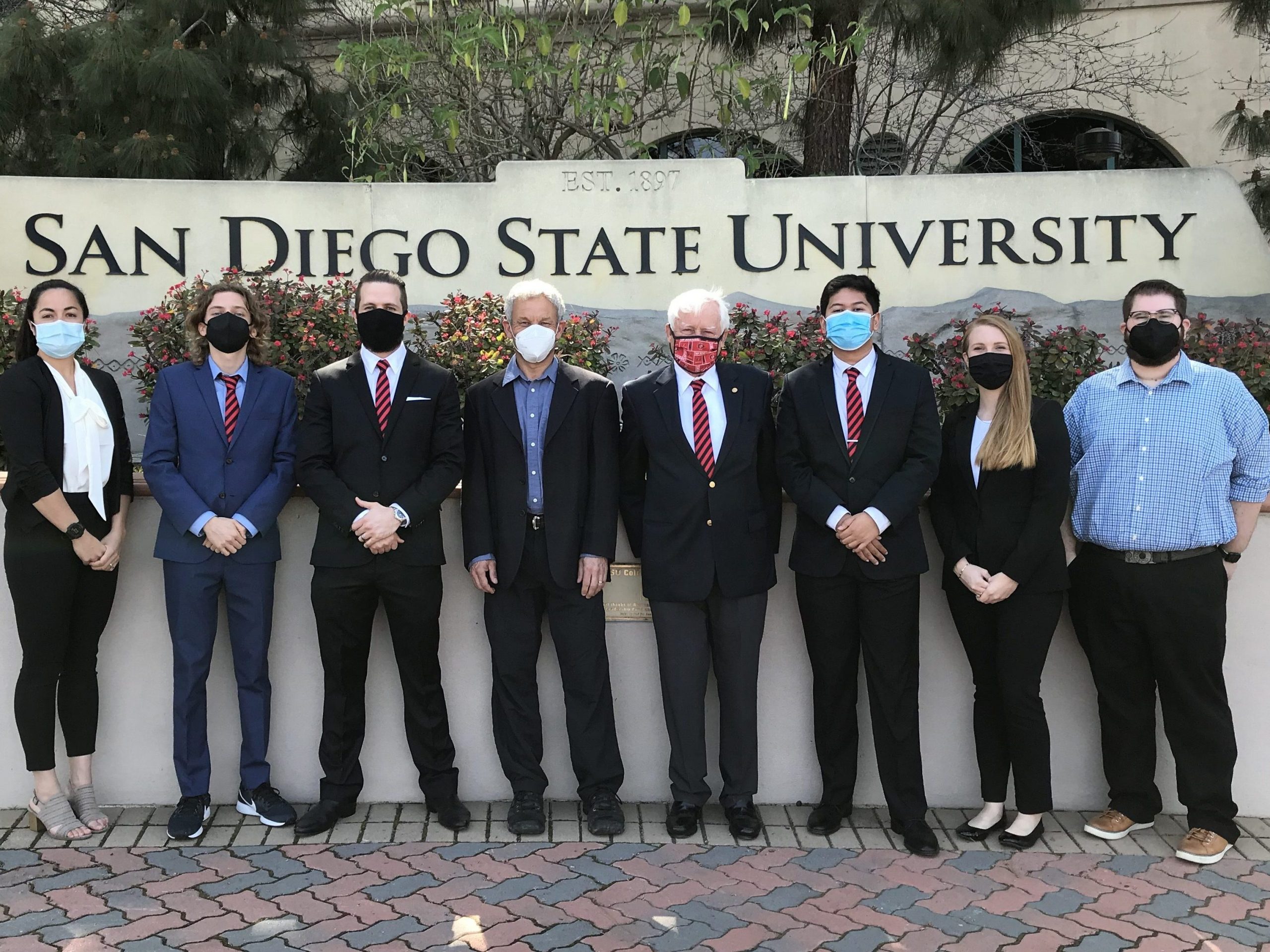 Jasmine Peach, Matthew Tinglof, Scott Smith, Kim Olsen, Kip Hering, Muhammad Fadilah, Hannah Carney, and Liam Schramm standing in front of the SDSU campus sign