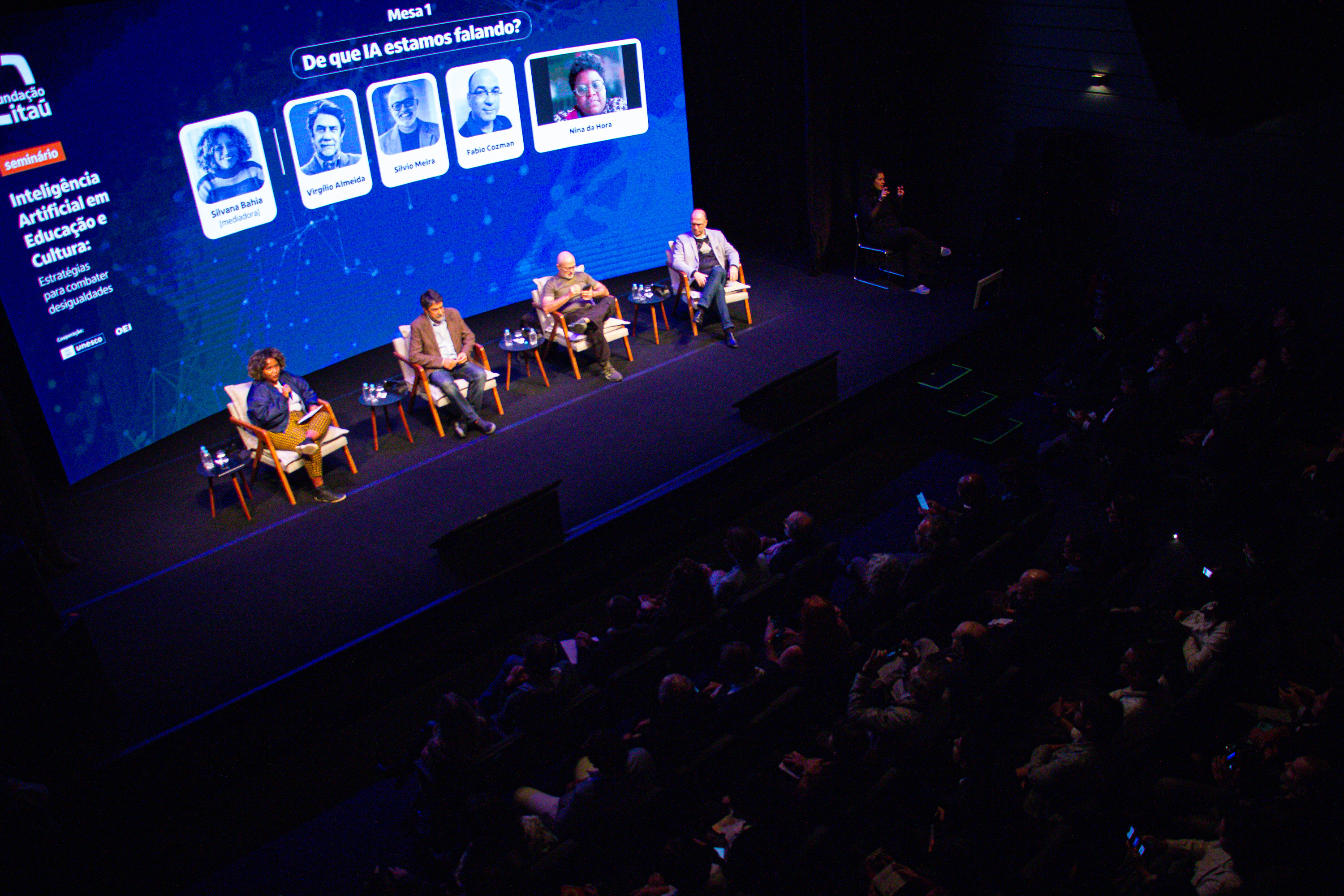 Quatro pessoas estão sentadas em um palco, com uma plateia a frente e atrás há um telão com o nome e foto de cada um, além da transmissão de vídeo de uma quinta pessoa participando online do evento.