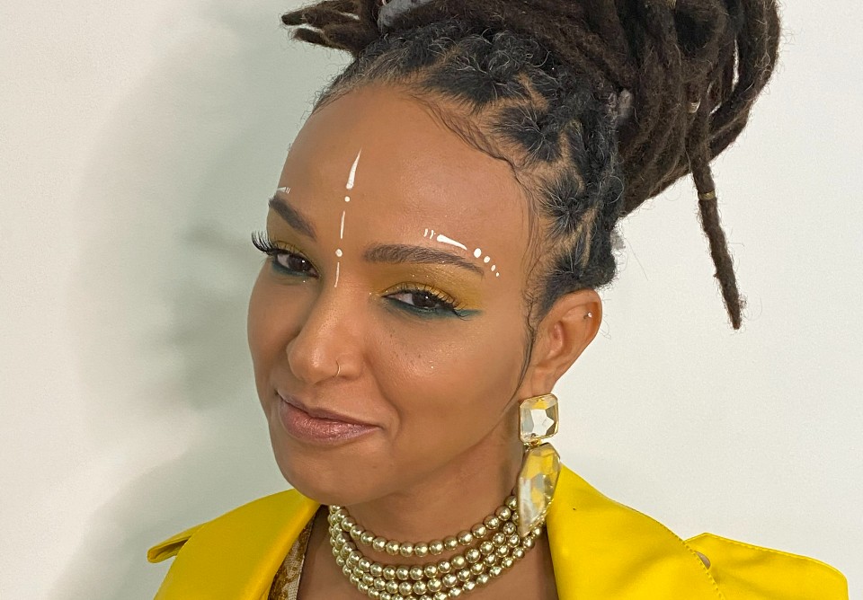 Silvanny é uma mulher negra com dreads. Ela olha para a câmera sorrindo e usa uma roupa amarela e um colar dourado.