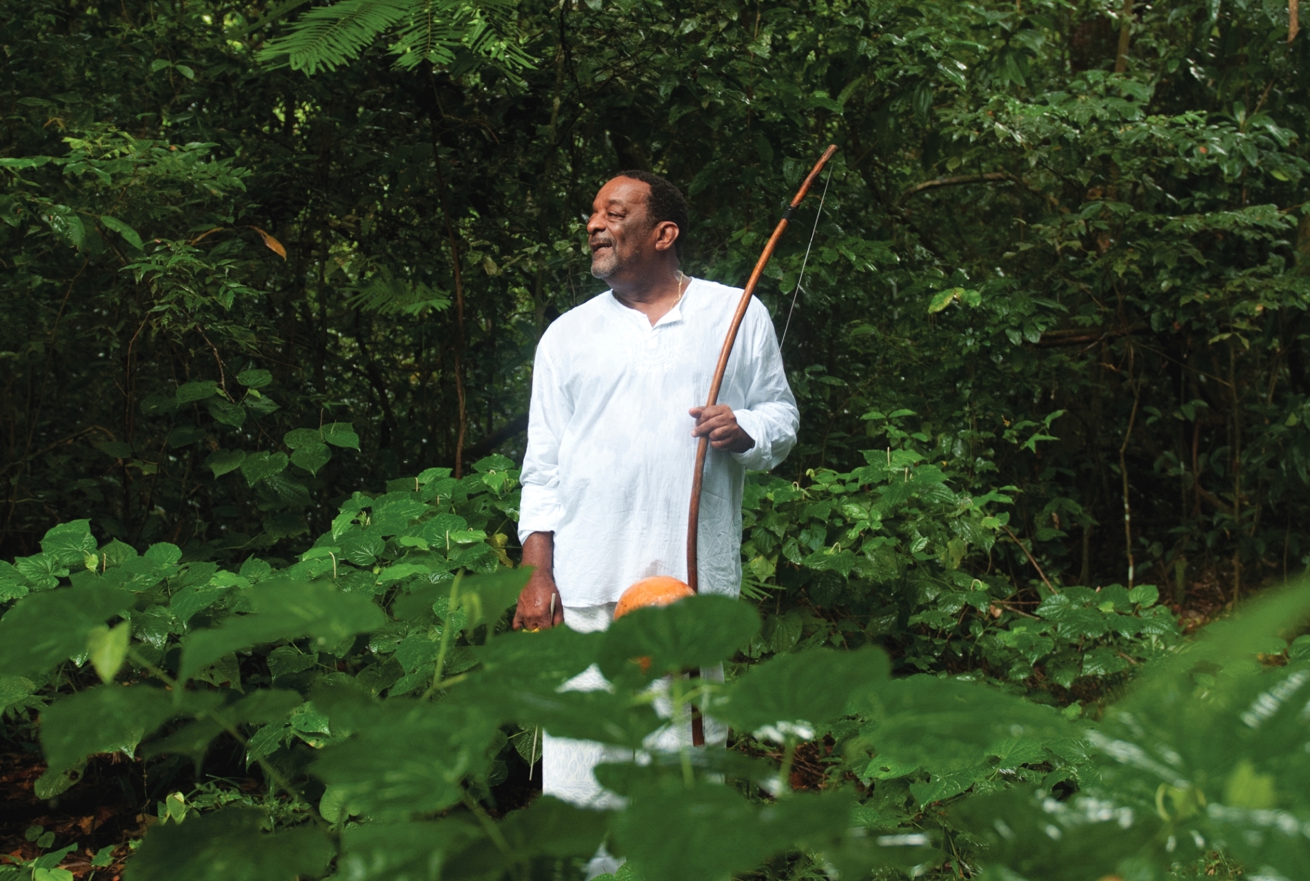 Fotografia colorida do músico Naná Vasconcelos. Ele é um homem negro, está vestido com uma roupa branca e segurando um berimbau. Está de pé no meio de uma área verde.