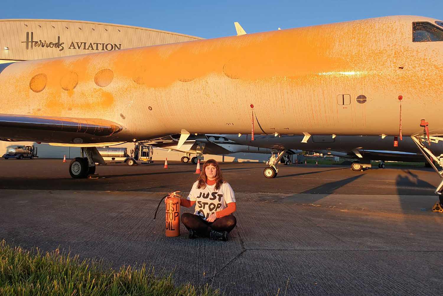 A handout picture released by the Just Stop Oil climate campaign group shows an activist sitting in front of a plane after spraying orange paint on private jets at Stansted Airport, northeast of London, on June 20, 2024. Climate activists gained access to a UK airport where Taylor Swift's luxury plane is stationed and sprayed private jets with orange paint, a day after Stonehenge was attacked.