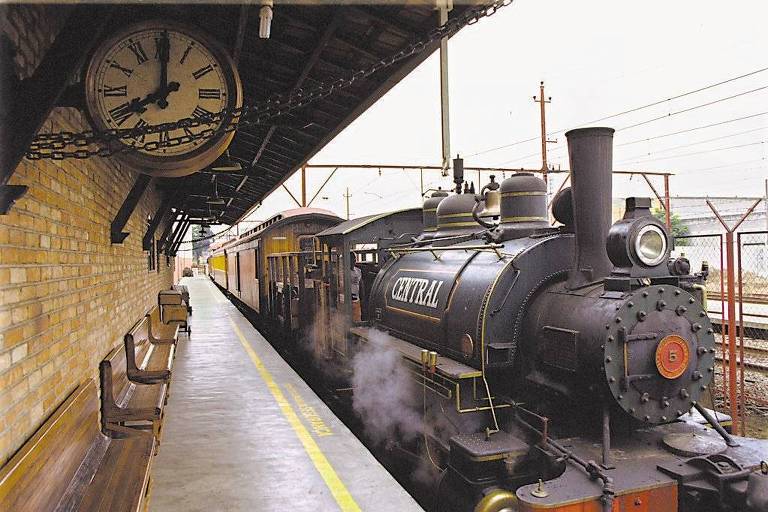 O trem est� parado em uma esta��o com cara de antiga, em S�o Paulo. � mostrada a plataforma vazia, e um rel�gio de ponteiro cl�ssico, com moldura de metal, est� suspenso no teto.