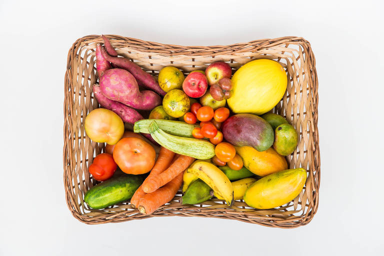 Foto com ângulo visto de cima de uma cesta de palha com futas e legumes meio tortos, com formatos fora dos padrões