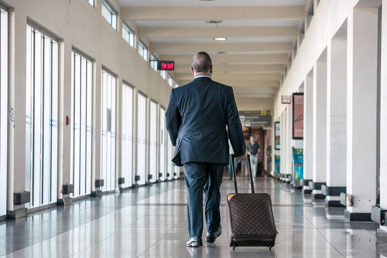 Homem de terno, de costas, puxa mala em corredor de aeroporto