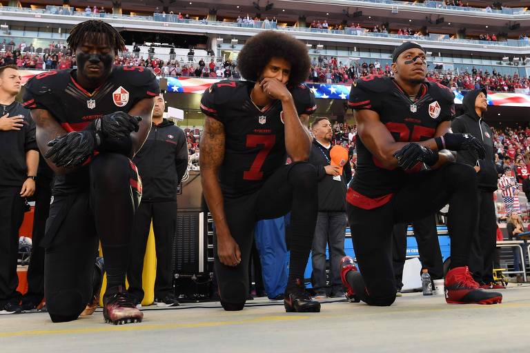 Eli Harold (à esq),  Colin Kaepernick (centro) e Eric Reid, do San Francisco 49ers, ajoelhados para protestar por justiça racial durante execução do hino nacional americano, em 2018