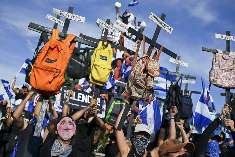 Grupo de dez manifestantes carregam cruzes pretas de madeira, cada uma delas com uma mochila pendurada e dois cartazes, com o nome da vítima e a cidade de onde ela veio. Entre eles há pessoas com bandeiras da Nicarágua e roupas com as cores do país.