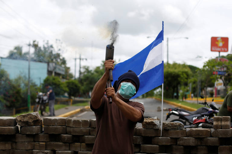 De camiseta marrom, máscara cirúrgica verde e gorro preto, manifestante segura o morteiro, feito com um cano preto e que solta fumaça antes de disparar. Imediatamente atrás dele está uma bandeira da Nicarágua que é sustentada por uma barricada feita de paralelepípedos. Atrás da barricada é possível ver outras duas pessoas à esquerda, também mascaradas, na calçada de uma rua asfaltadas com os meios-fios pintados de amarelo.