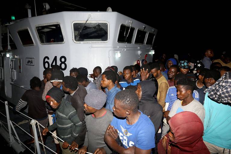 Cerca de 40 homens se amontoam de pé em convés de barco; à direita acima, a torre de controle