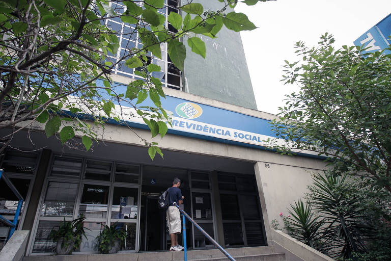 Foto mostra a fachada de uma agência da Previdência Social. O prédio é cinza com uma placa comprida azul com o símbolo do INSS ao lado dos dizeres Previdência Social. Há galhos de árvores com folhas verdes nas duas laterais da foto. Na porta da agência, tem um homem de mochila parado em pé.