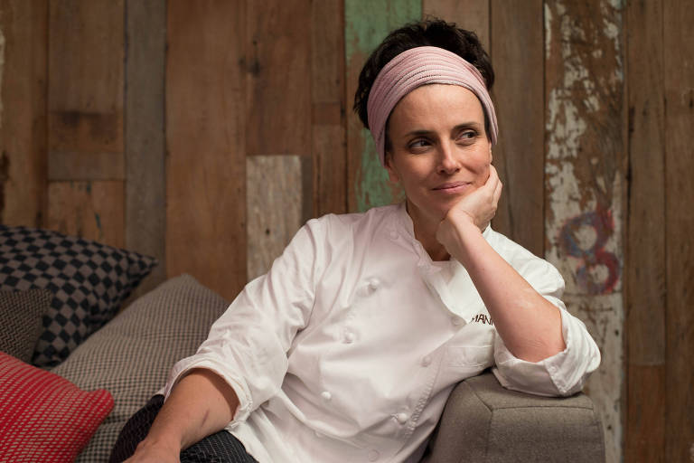 A chef Helena Rizzo sentada em um sofá com uniforme branco e cabelo preso por uma faixa, apoiada em um dos braços; ao fundo, parede de madeira  