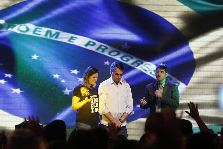 Um telão exibe uma enorme bandeira do Brasil. Diante dela, o presidente e a primeira-dama oram de cabeça abaixada, enquanto um pasto fala ao microfone