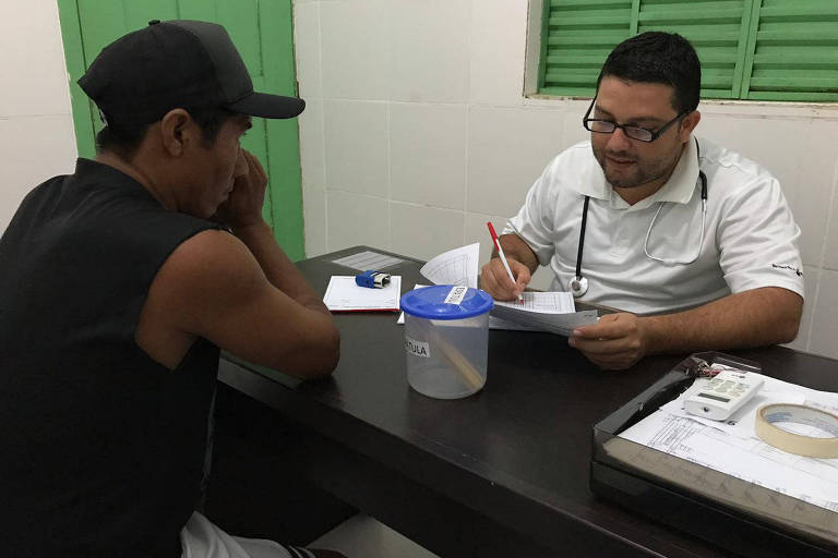 Imagem mostra homem indígena vestindo camiseta preta e usando boné sentado em mesa de frente a médico com roupas brancas, que segura um papel e porta um estetoscópio sobre o pescoço