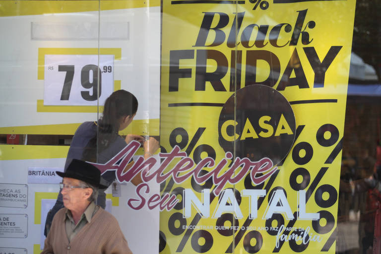 Vitrine de loja na cidade de Campinas que diz Antecipe seu Natal, com senhor vestido de pullover bege, chapéu preto e óculos a frente. Ao fundo, funcionária ajusta cartaz com o escrito Black Friday Casa, na cor amarela.