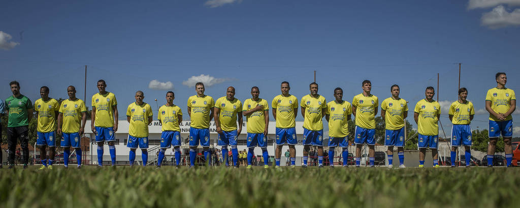 Homens com calção azul e camiseta amarela perfilados em gramado 