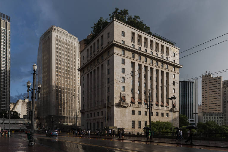 A imagem mostra uma vista do Edifício Matarazzo, sede da Prefeitura de São Paulo. O prédio tem uma fachada clara e janelas retangulares. O edifício está cercado por outros prédios e o céu está nublado ao fundo. Algumas pessoas estão caminhando na calçada em frente ao edifício.