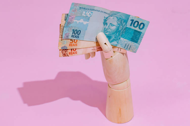 Foto mostra um fundo rosa com uma mão articulada de madeira segurando notas de 100, 20 e dez reais.